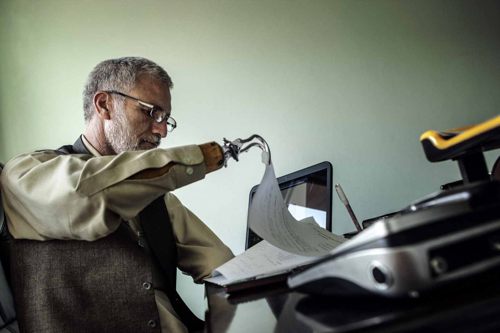 Pildi vasakus servas on halli siilisoengu ja prillidega mees, seljas kollakasvalge triiksärk ja tume vest. Mees on küljega vaataja poole, näoga paremale. Mehe parem käsivars on tõstetud, selle otsas olev metallist traatsilmuse moodi protees tõstab tema ees oleval kaldlaual paberipatsaka pealmist lehte. Mehe vasakul käel paistab laual arvutiekraan, esiplaanil on ähmaselt, fookusest väljas, hõbehall seadeldis, mille vaataja poole pööratud tagaküljes on ümmargune pistik. Pildi taust on ühtlaselt kollakashall.