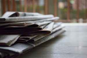 Siledal pinnal on kokku volditud ajalehtede virn, taustal on näha ähmased vertikaalsed piirded ja haljastus. Tähelepanu on suunatud ajalehtede servadele esiplaanil.