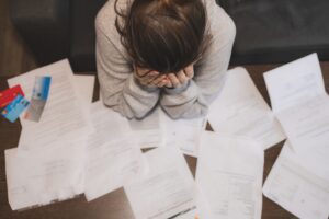 Inimene istub laua taga, pea kätes, ümbritsetud hajutatud paberitest ja krediitkaartidest, mis viitab stressile või ülekoormatusele - võimalik, et see on seotud rahaasjade, arvete, hooldajatoetuse või ravikindlustuse probleemidega.