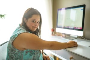 Noor naine, kellel on Downi sündroom, istub arvuti kõrval laua taga, vaatab üle õla ja naeratab vaikselt. Ta kannab helesinist mustrilist kleiti ja ruumis on loomulikku valgust.