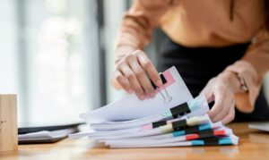 Inimene korrastab puidust laual köiteklambritega kokku klammerdatud dokumendipakke, mille sektsioonid on tähistatud värviliste sedelitega.