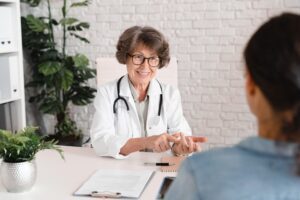 Sõbralik prillidega naisarst istub oma töölaua taga ja selgitab midagi patsiendile. Arstil on seljas valge kittel ja kaelas stetoskoop. Patsienti on näha selja tagant, tal on seljas helesinine jakk. Ruum on valgusküllane, taustal on telliskivisein ja toataimed.