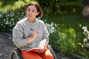 Lühikeste tumedate juustega naine istub ratastoolis päikesepaistelises aias või pargis. Tal on seljas hall pusa ja jalas oranžid püksid. Naine hoiab süles avatud märkmikku ning paremas käes pastapliiatsit.