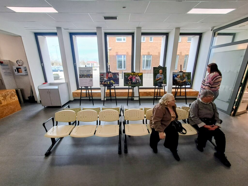Omastehooldajate fotonäituse avamine Ahtme tervisemajas: kolm külastajat vaatavad statiividel väljapandud portreefotosid akna ees, taustal nähtav silt tekstiga "Omastehooldaja – tavaline eriline imeline inimene."