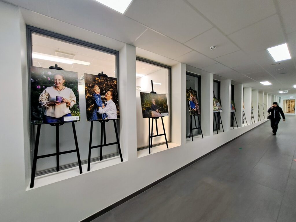Omastehooldajate fotonäitus Ahtme tervisemaja koridoris: rida portreefotosid statiividel aknaavade ees, esiplaanil naine kohvitassi ja aiaõite taustal, kõrval foto emast ja lapsest.