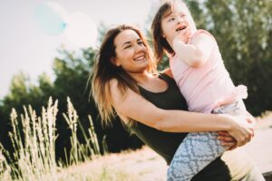 Naeratav naine hoiab käte vahel väikest tüdrukut, kellel paistavad Downi sündroomi tunnusjooned, päikeselisel niidul, taustal puud ja õhus hõljuvad sinised õhupallid.