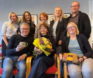 Kaheksa kolleegi poseerivad grupipildil kontoris. Ees istub kolm inimest — üks hoiab kruusi, üks kollaste ja roosade lillede kimpu, üks karbis mesikooki; taga seisab viis inimest.