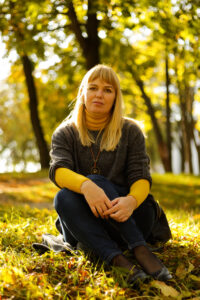 Esiplaanil istumas tumedates riietes heledapäine naisterahvas vaatamas otse kaamerasse, Tema taustal sügisene loodus rohekates-kollakates toonides.
